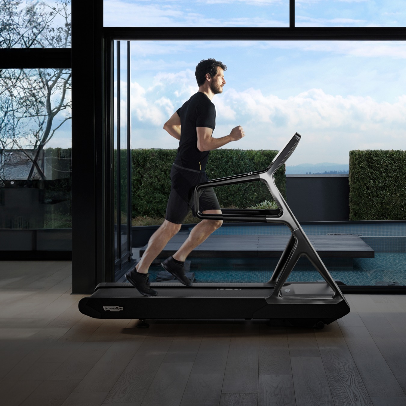 A man wearingblack shorts and tshirt running on a luxury treadmill with shiny metal finishes (Technogym Run Personal) in a luminous apartment with woodboard flooring, a small pool can be seen on the terrace right outside